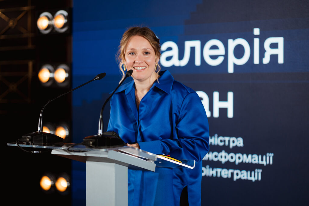Valeriia Ionan speaks at the Ukraine Digital Transformation Activity launch event. A large screen behind her shows her name in Ukrainian.
