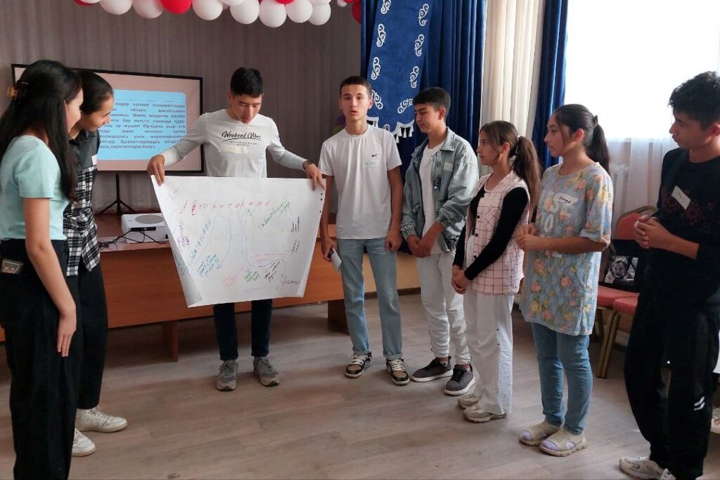 Seven students stand in a semi-circle. One holds up a large sheet of paper with writing on it.